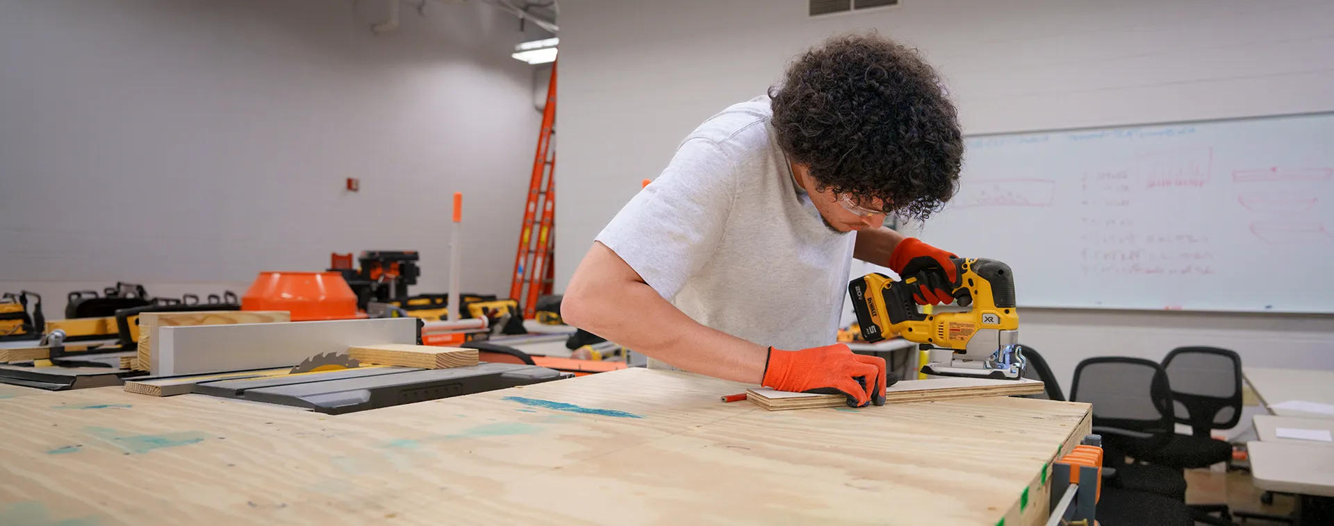 Person using a jigsaw on wood at a workbench in a workshop with tools, ladder, and whiteboard in background