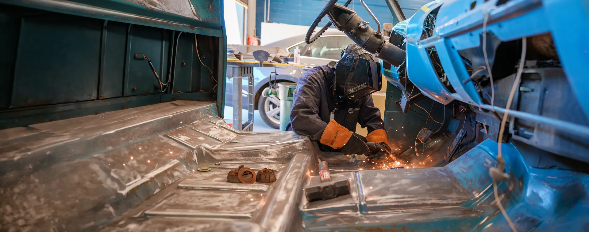 Technician welding inside stripped vehicle body, sparks flying across metal floor panels and tools nearby.