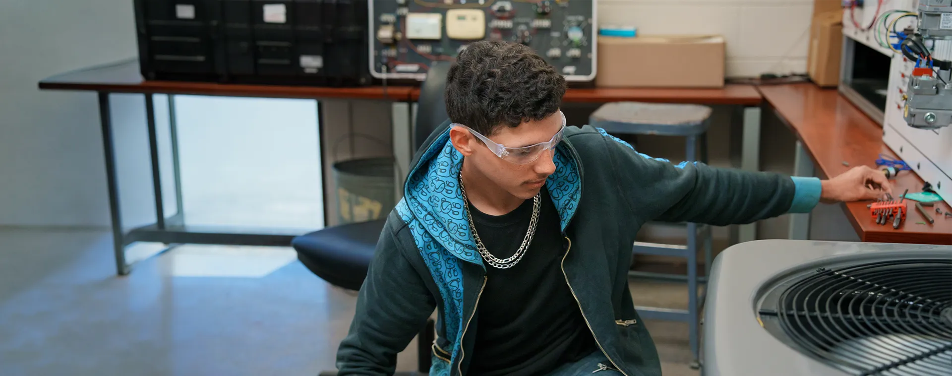 Person wearing safety glasses sits beside HVAC unit, reaching toward components with tools on nearby desk.