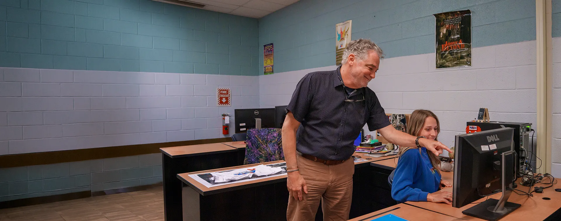 Instructor pointing at computer screen while student works at desktop in classroom with posters and desks