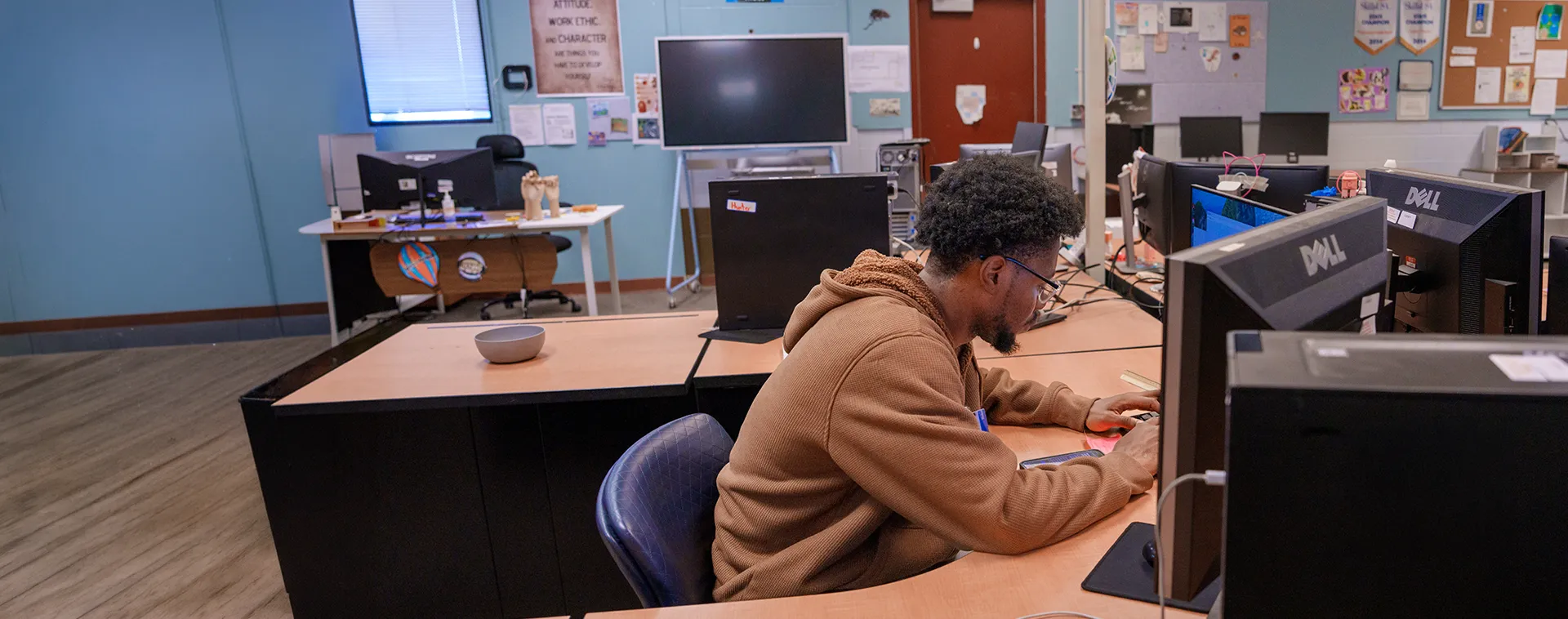 Person seated at desktop computer in classroom lab with multiple monitors, desks, and bulletin board on wall