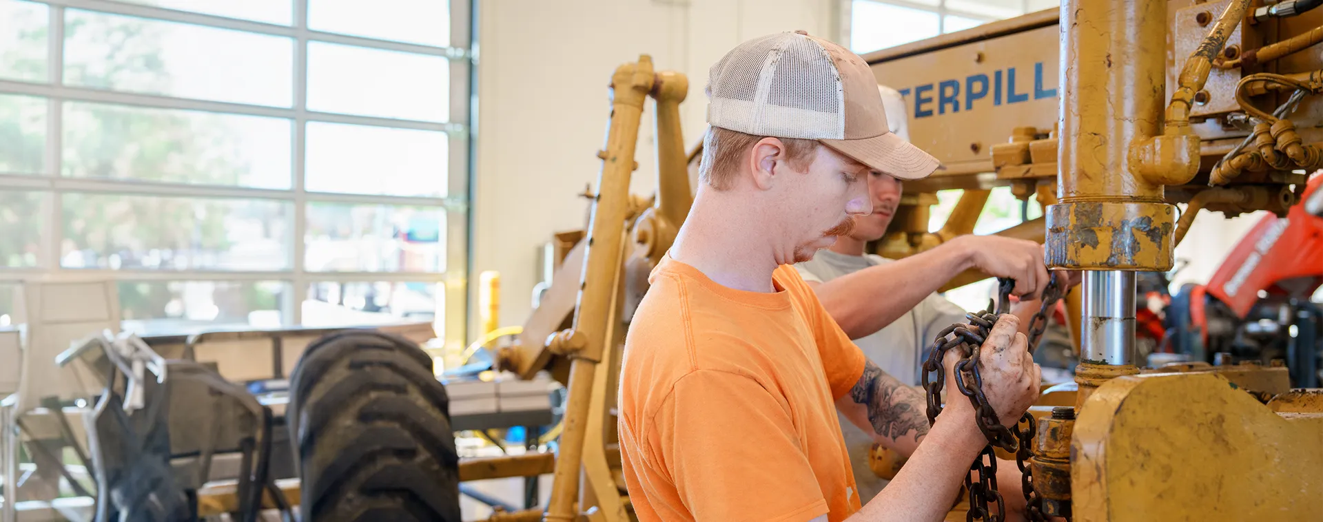 Two people working on a Caterpillar machine, handling a chain in a bright workshop with large windows and equipment