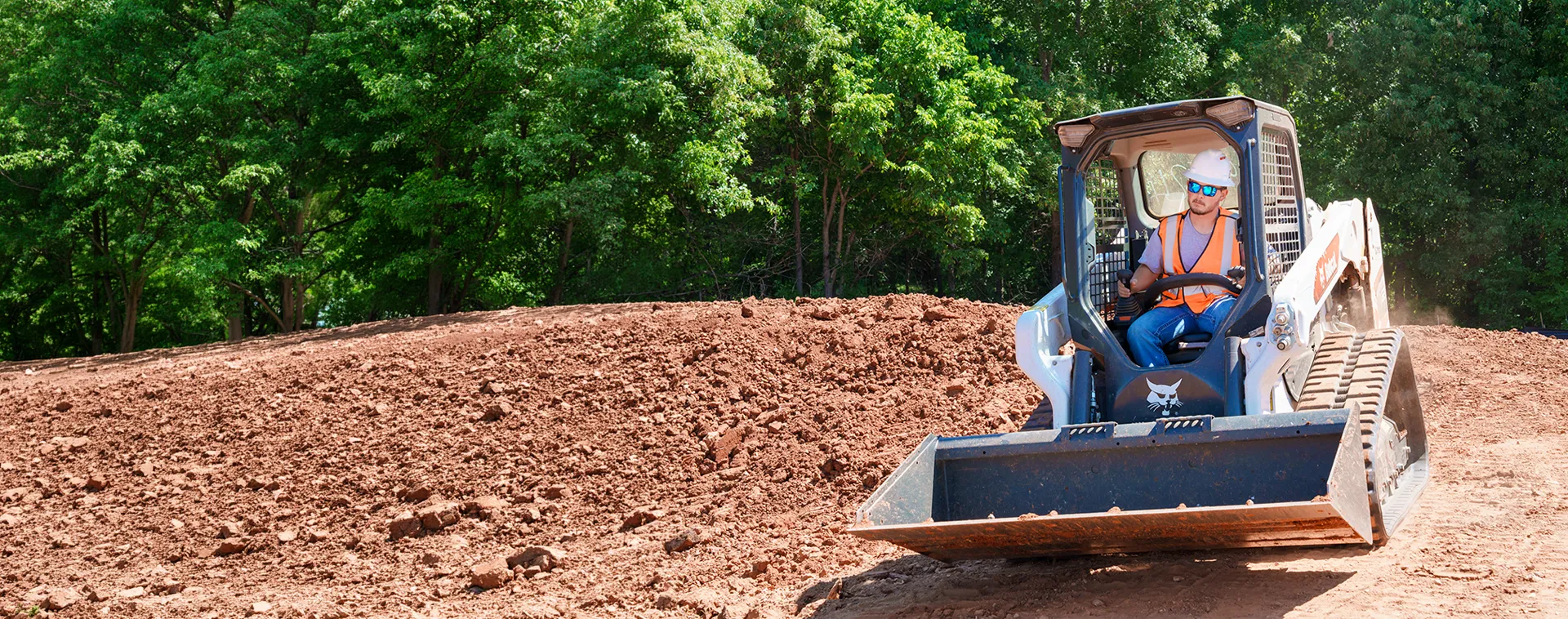 Person operating a compact track loader on dirt field with trees in background