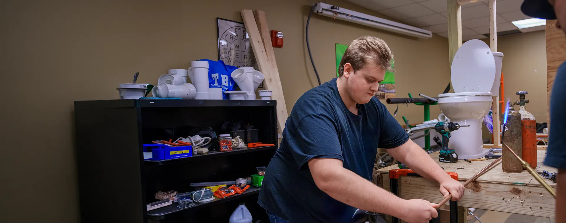 Person bending copper pipe at workbench with tools, PVC fittings, and toilet fixture in workshop