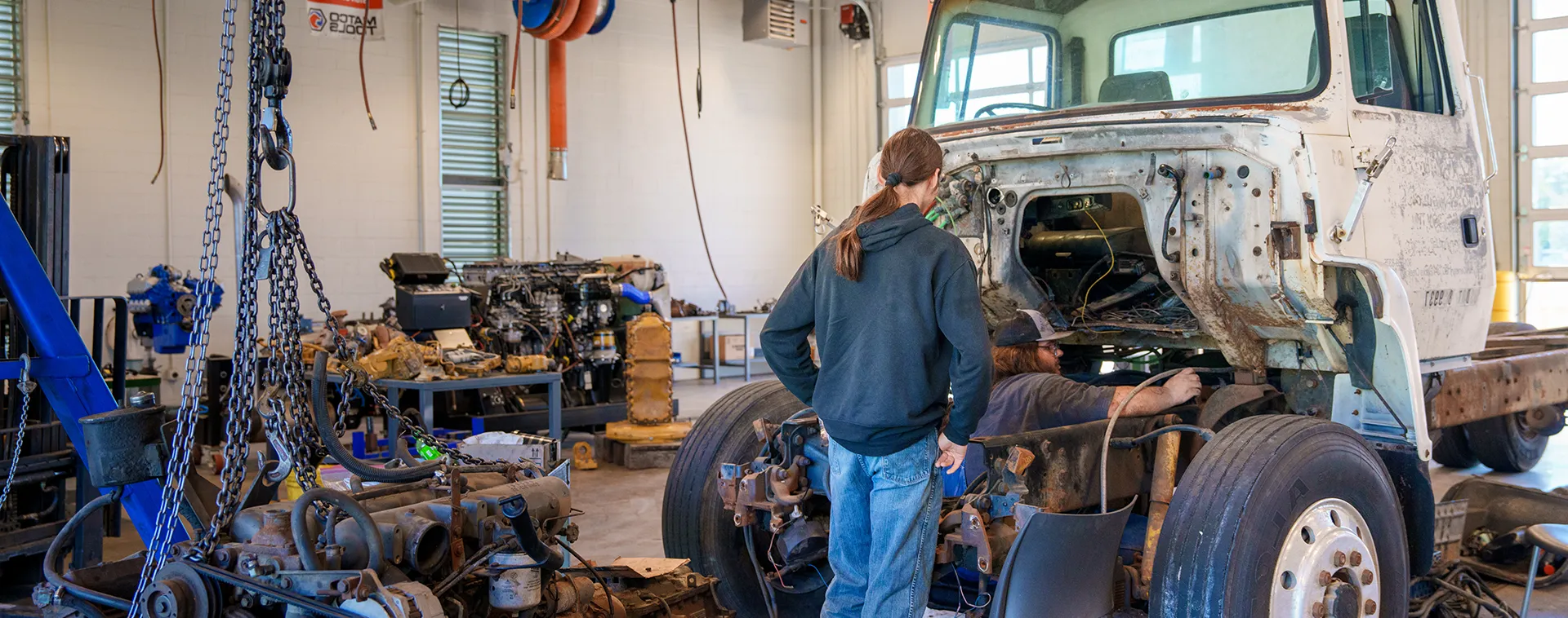 Two people work on exposed engine of large truck inside shop with chains, tools, and parts on nearby tables.
