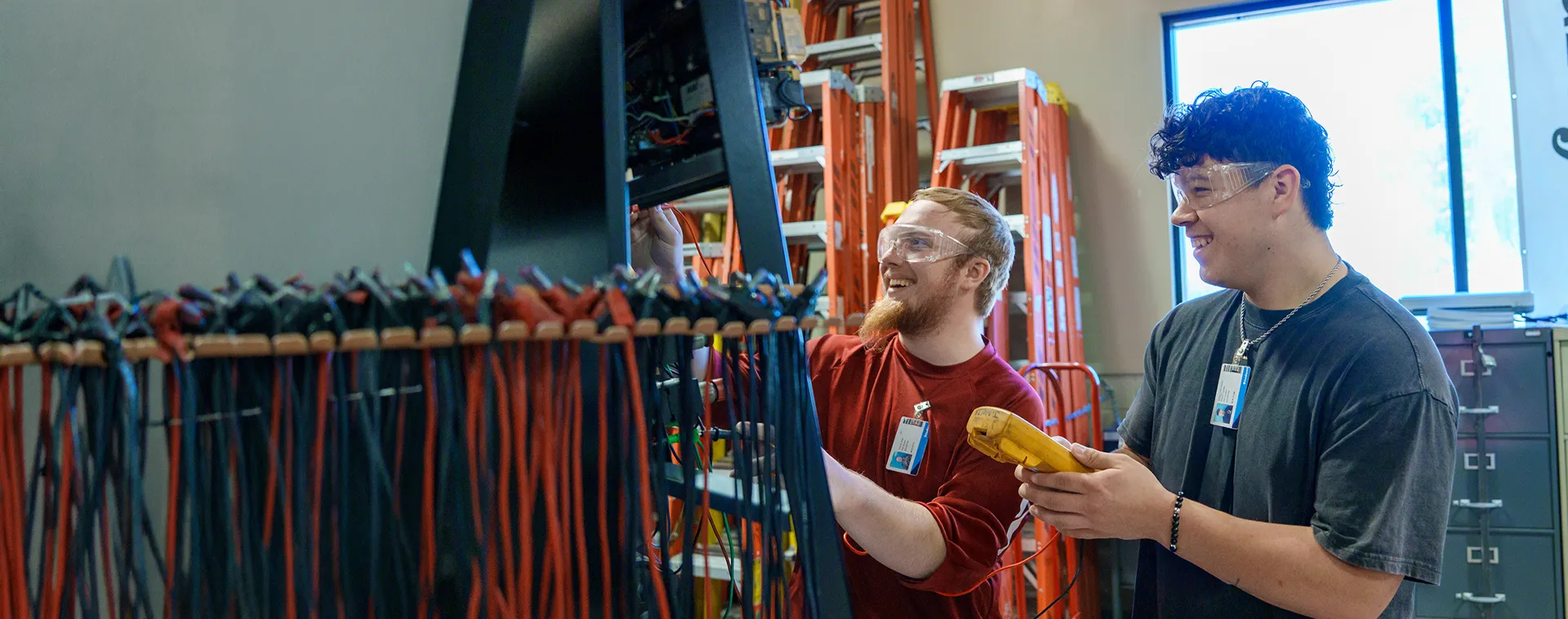 Two people wearing safety glasses connect wires on a training panel with bundled cables and ladders behind.