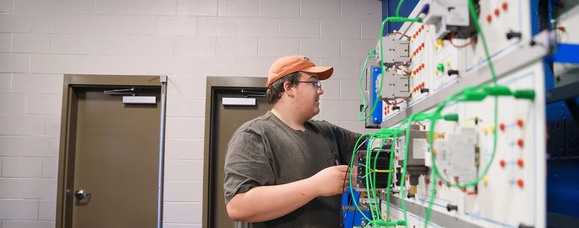 Person in cap connects green wires on electrical control panel with switches and modules mounted on wall.