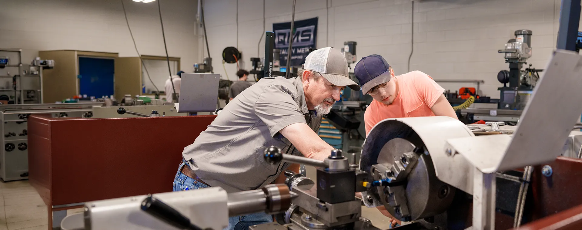 Instructor and student operate metal lathe machine in workshop with other equipment and workstations in background.