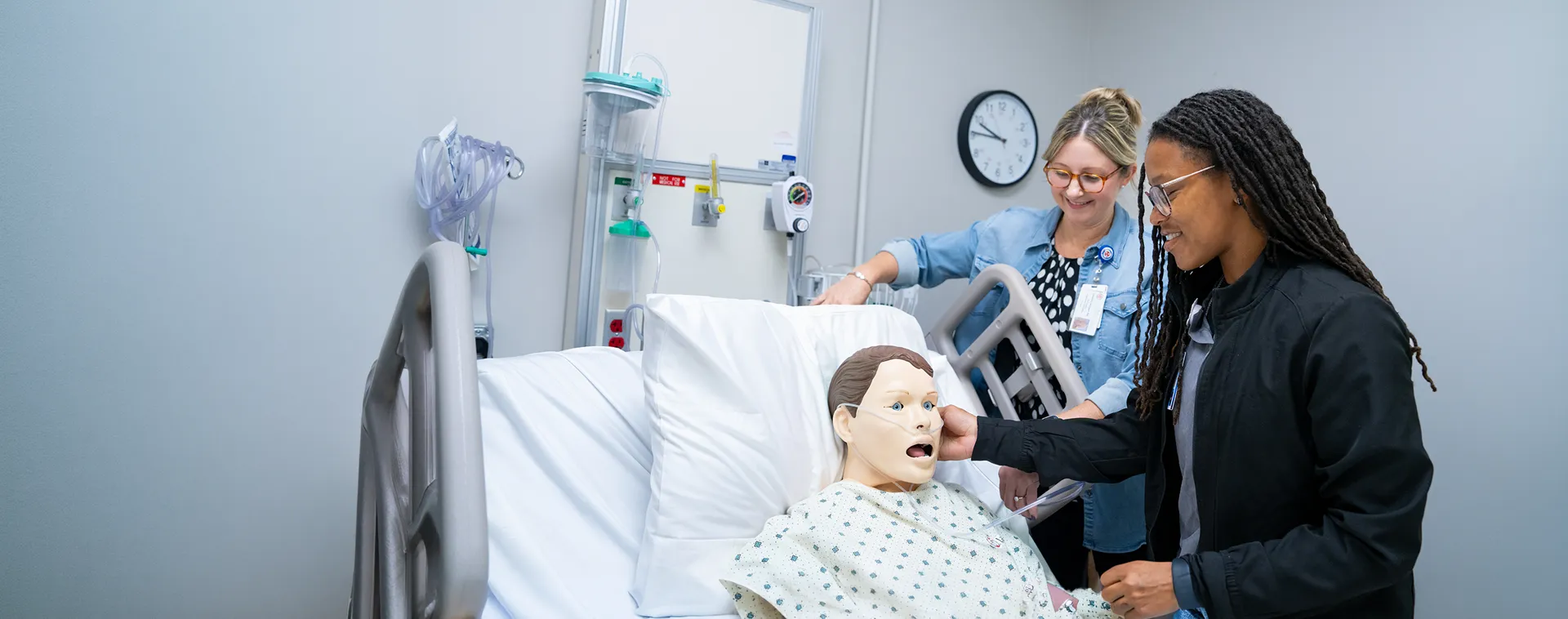 Two people adjust a patient simulator in hospital bed while another observes equipment on wall and monitor.