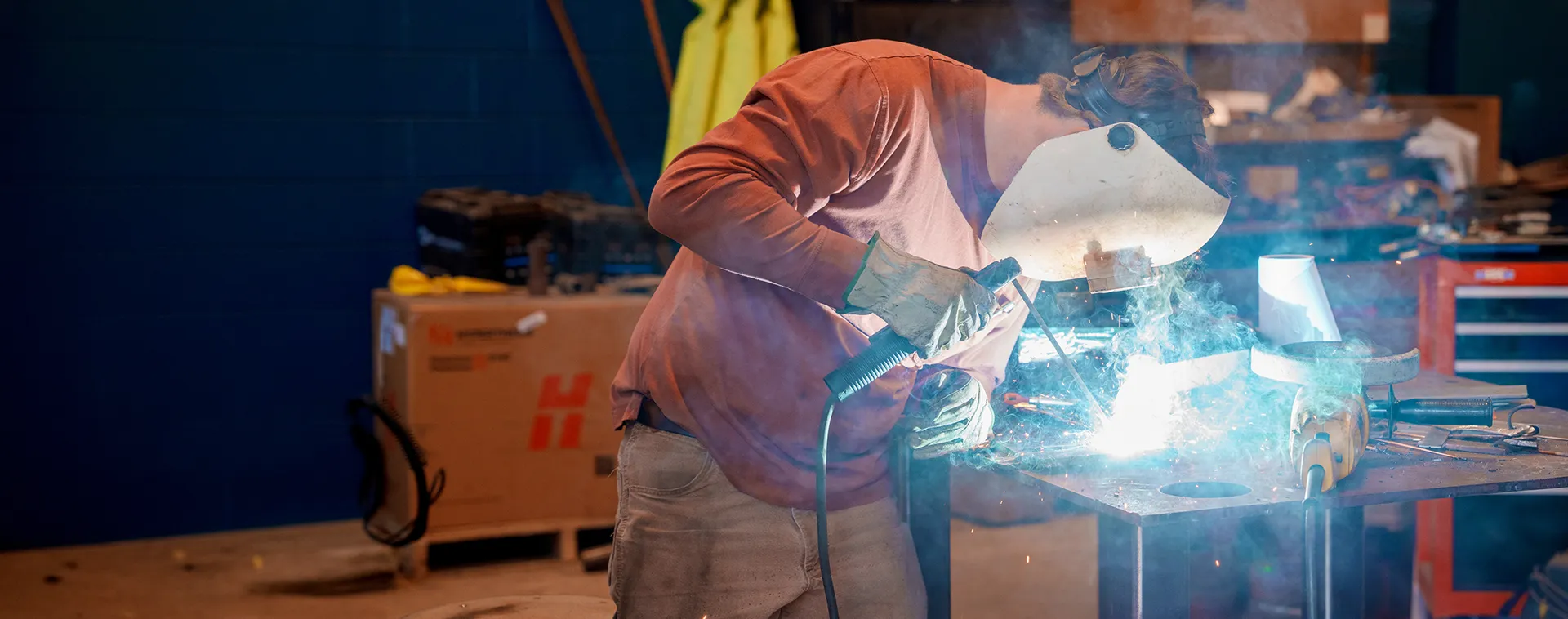 Person wearing welding helmet and gloves uses torch on metal, bright arc light and sparks on workbench in shop.
