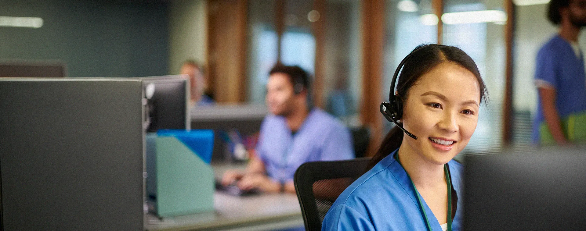 Person wearing headset and scrubs works at computer in office with other staff and monitors in background.