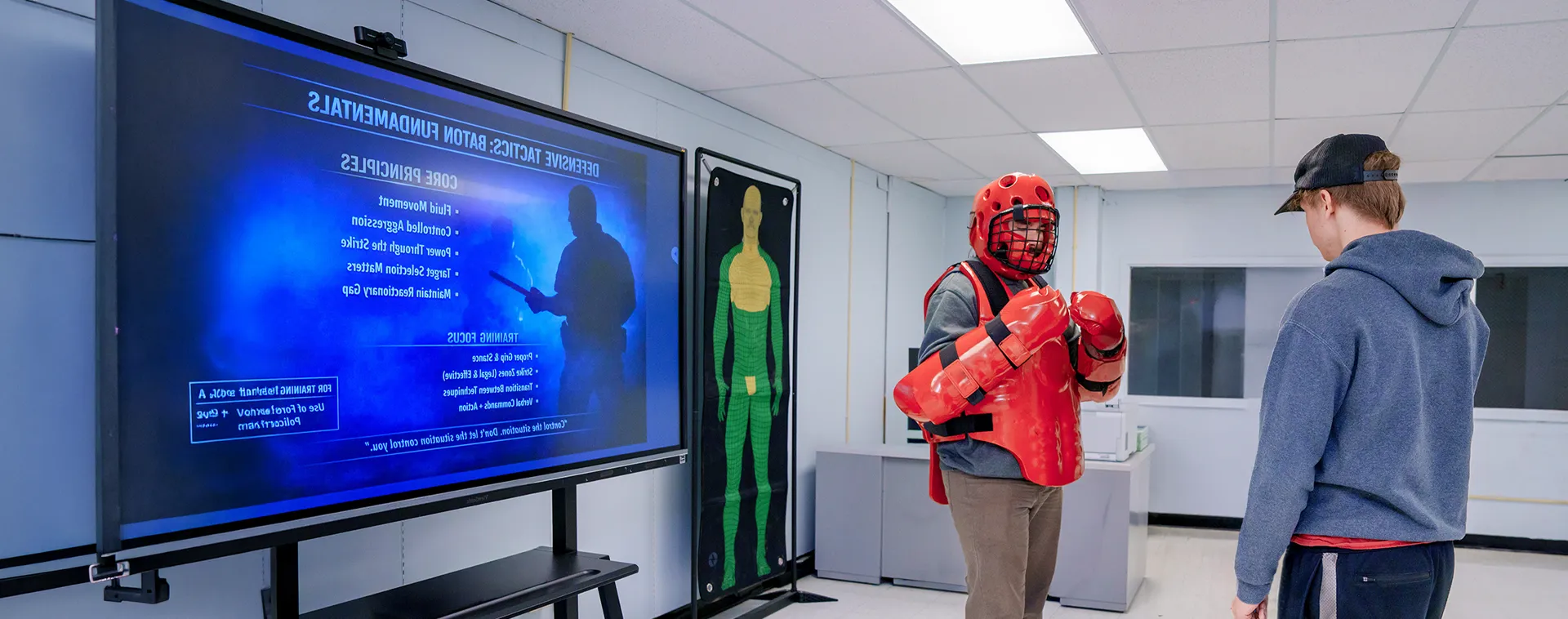 Instructor guiding two students in red protective gear during defensive training activity in classroom with wall posters and display screen