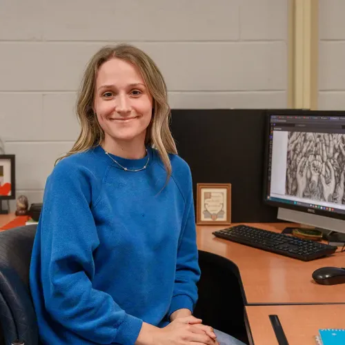 Person seated at desk smiling toward camera beside computer showing digital artwork on monitor