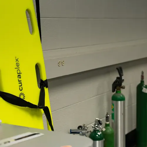 Yellow spine board labeled &ldquo;curaplex&rdquo; leaning near oxygen tanks and medical equipment on shelf.