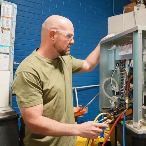 Technician wearing safety glasses adjusts valves and gauges on open HVAC unit against blue wall.