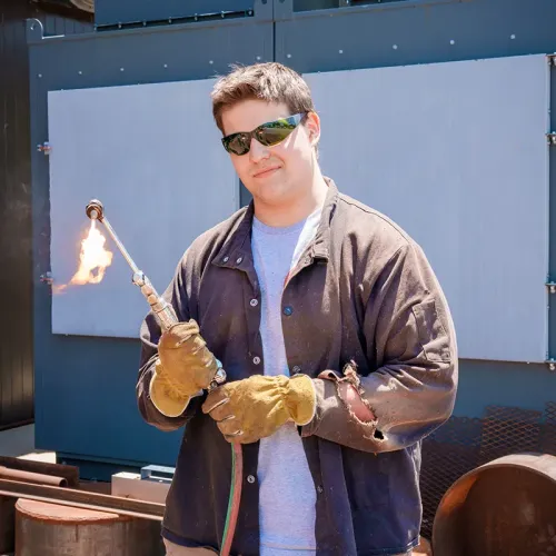 Person wearing gloves and safety glasses holds lit torch flame outdoors near metal equipment and panels.