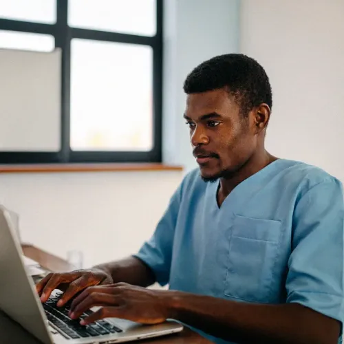 Person in scrubs types on laptop at desk with window and wall behind.