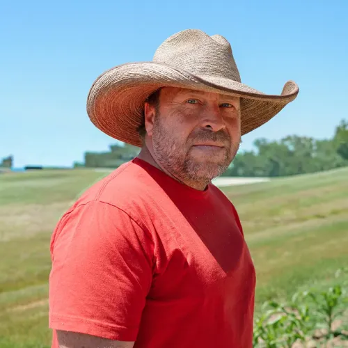 Person wearing wide-brim hat and red shirt stands in grassy field with crops and trees in background.