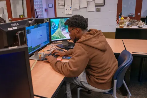 Person using dual monitors and keyboard at desk in computer lab with papers and equipment on walls