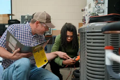 Instructor and student kneel by HVAC system using multimeter while reviewing an energy guide booklet.