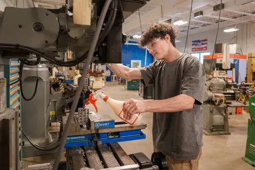 Person uses spray bottle to clean drill press table in machine shop filled with industrial tools.