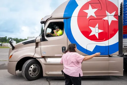 Person stands beside semi truck cab while driver sits inside; large TCAT logo displayed on truck door.