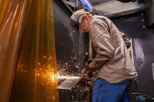 Person uses angle grinder on metal inside welding booth, sparks flying against protective screen.