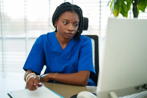 Person in blue scrubs writes on paper while looking at desktop computer beside window and indoor plant.